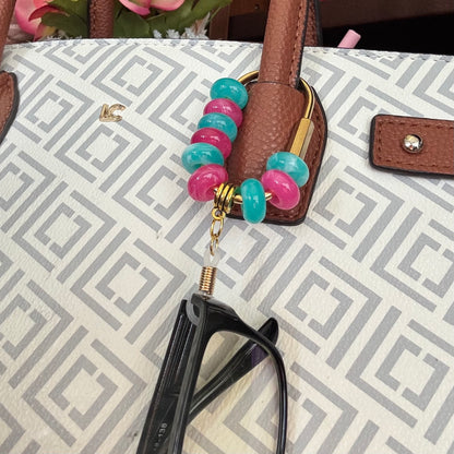 A close-up of a handbag with a geometric pattern in beige and gray, featuring a brown leather handle adorned with a beaded keychain in pink and teal with gold hardware. A pair of black eyeglasses rests below the handle.